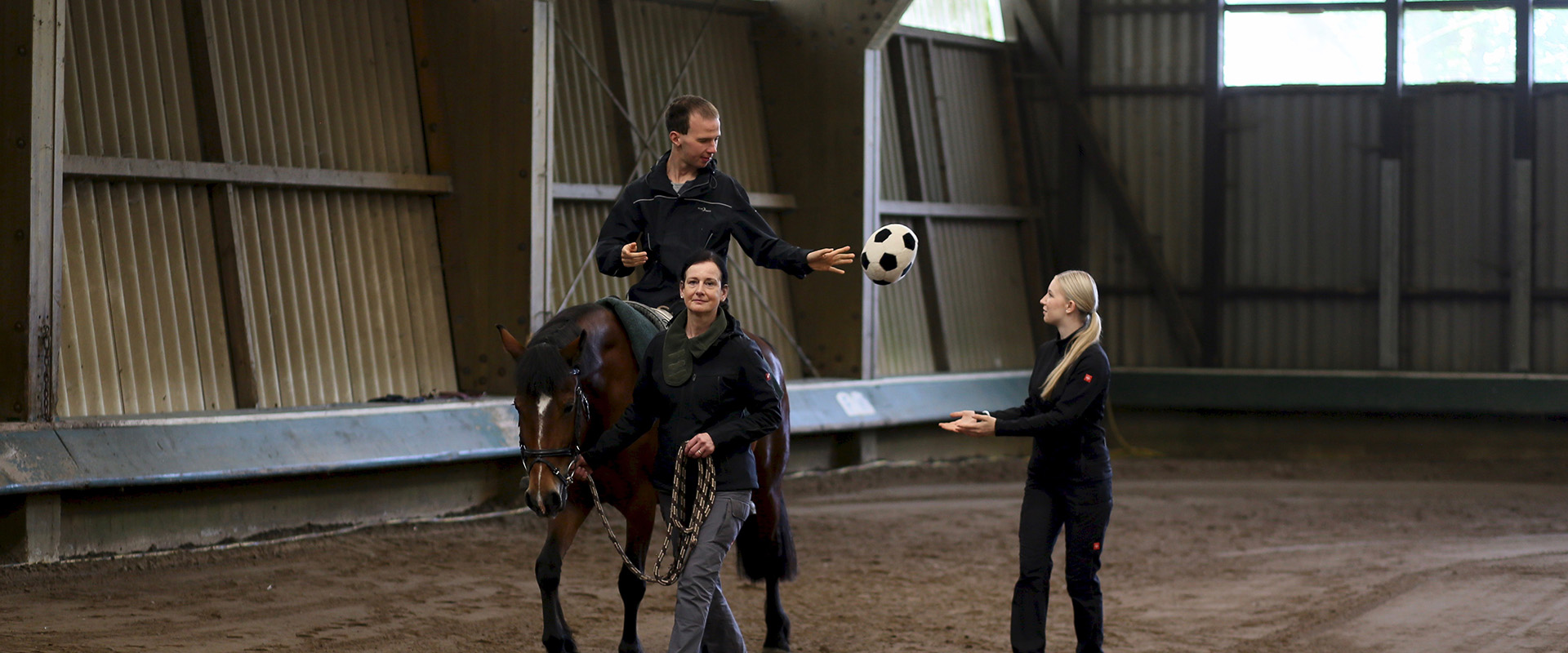Ein körperlich beeinträchtigter Mann sitzt auf einem Pferd das geführt wird und wirft einen Ball zu einer Frau